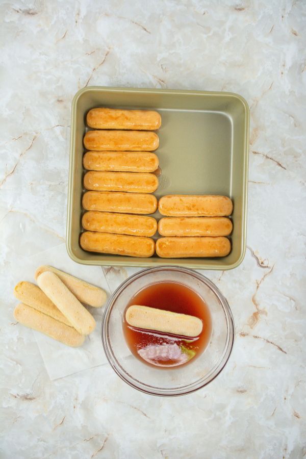 Delicate ladyfinger biscuits are being dipped into a vibrant red strawberry mixture beside a baking dish, meticulously layered for the perfect No Bake Strawberry Tiramisu.