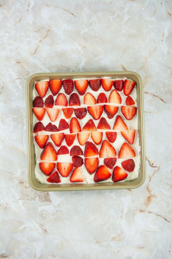 A square pan displays a No Bake Strawberry Tiramisu, with layered sliced strawberries meticulously arranged atop a creamy dessert in neat rows.