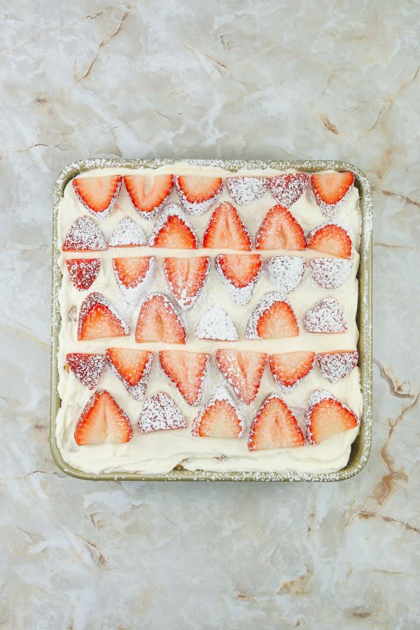 Square cake with sliced strawberries and powdered sugar on top, this no-bake creation mimics a strawberry tiramisu, elegantly set on a marbled surface.